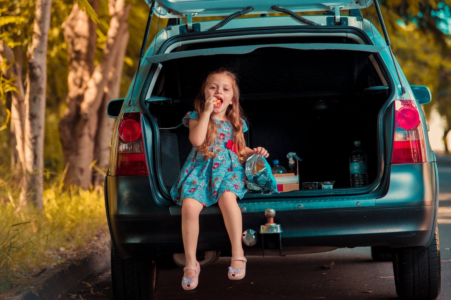 a young girl in blue floral dress sitting on the car trunk while eating food