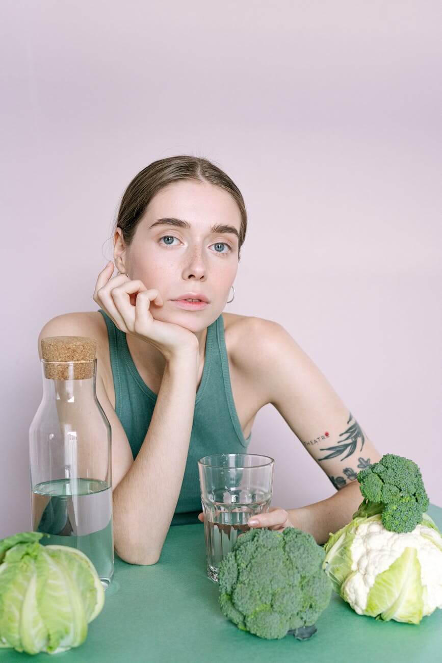 woman in green tank top sitting beside table with drinking glass and fresh vegetables